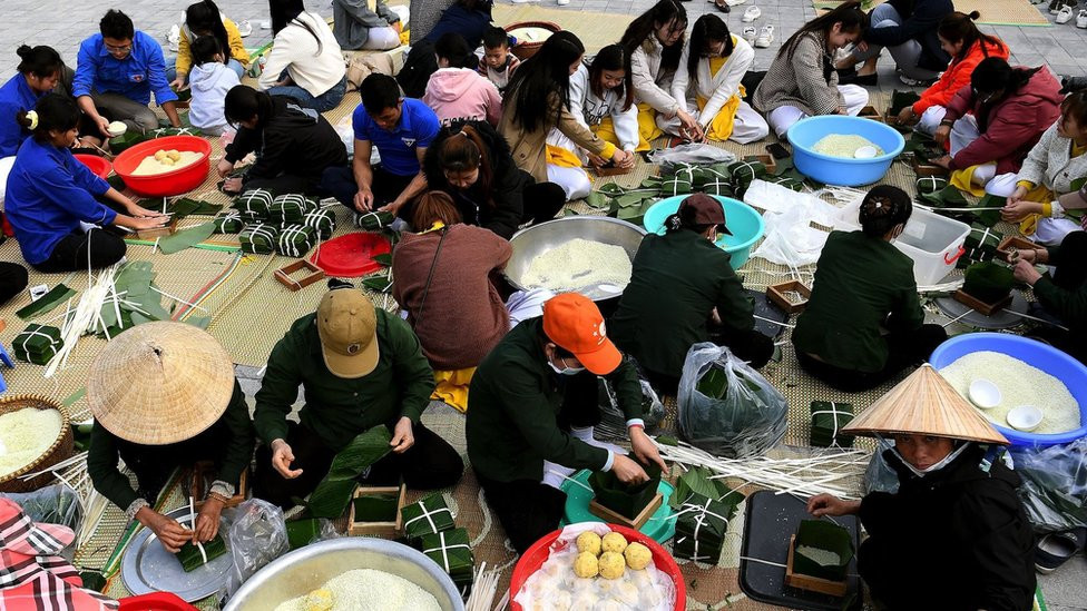 A large group of people sitting on mats making rice cakes