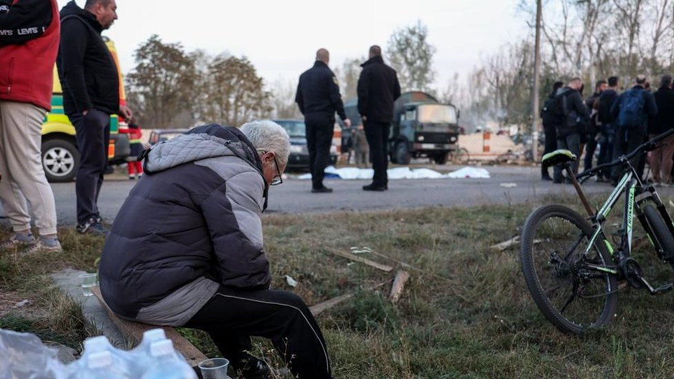 Local residents at the site of a military strike in the village of Hroza, northeastern Ukraine on 5 October 2023. A man is seen in the foreground sitting on a concrete ledge amid a patch of grass.