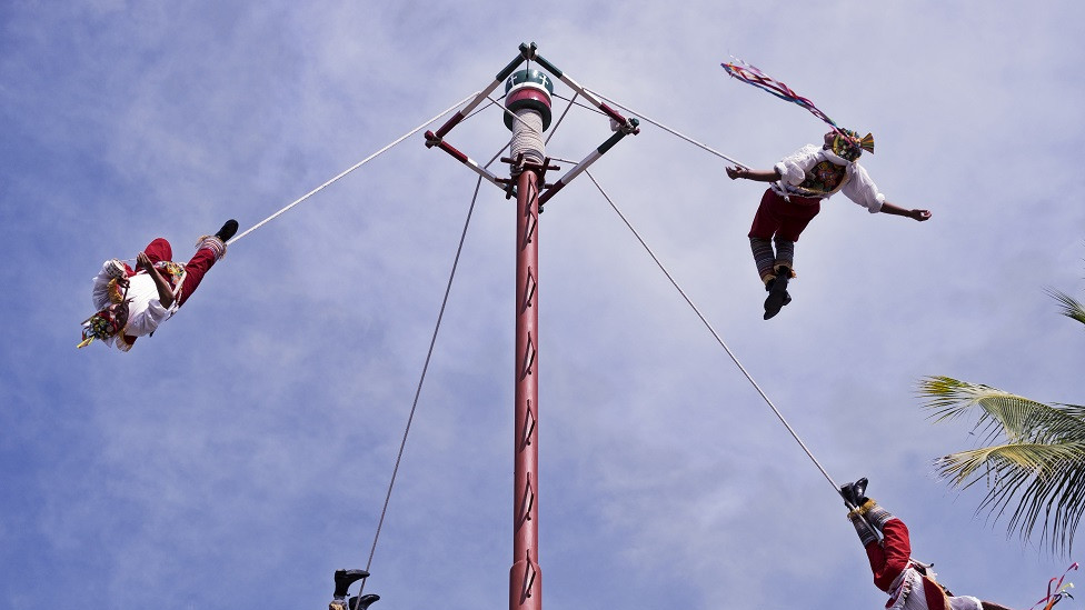 Four men seeming to dance in the air, attached by ropes to a very tall pole