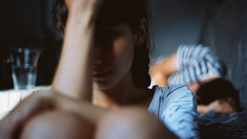 Annoyed woman sitting by the bed with snoring boyfriend at home - stock photo