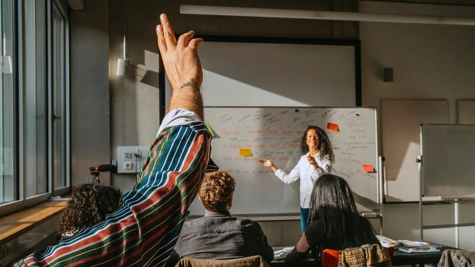 Žena na času sa odraslim studentima među kojima jedan drži podignutu ruku