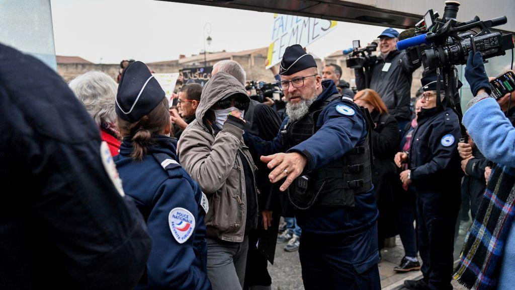 Jedan od optuženika stiže u sudnicu pokrivenog lica sa kapuljačom na glavi okružen policajcima