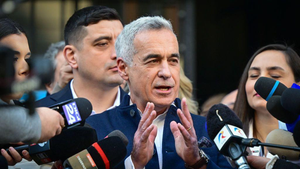 Muškarac u plavom odelu gestikulira rukama pred mikrofonima