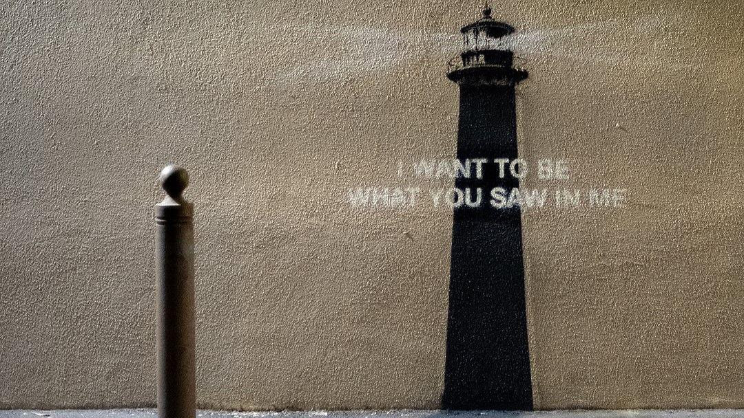 
                    A photo of the Banksy piece of art. A metal bollard is in the foreground on the pavement, while a painting of a lighthouse is on the wall superimposed with a quote "I wan