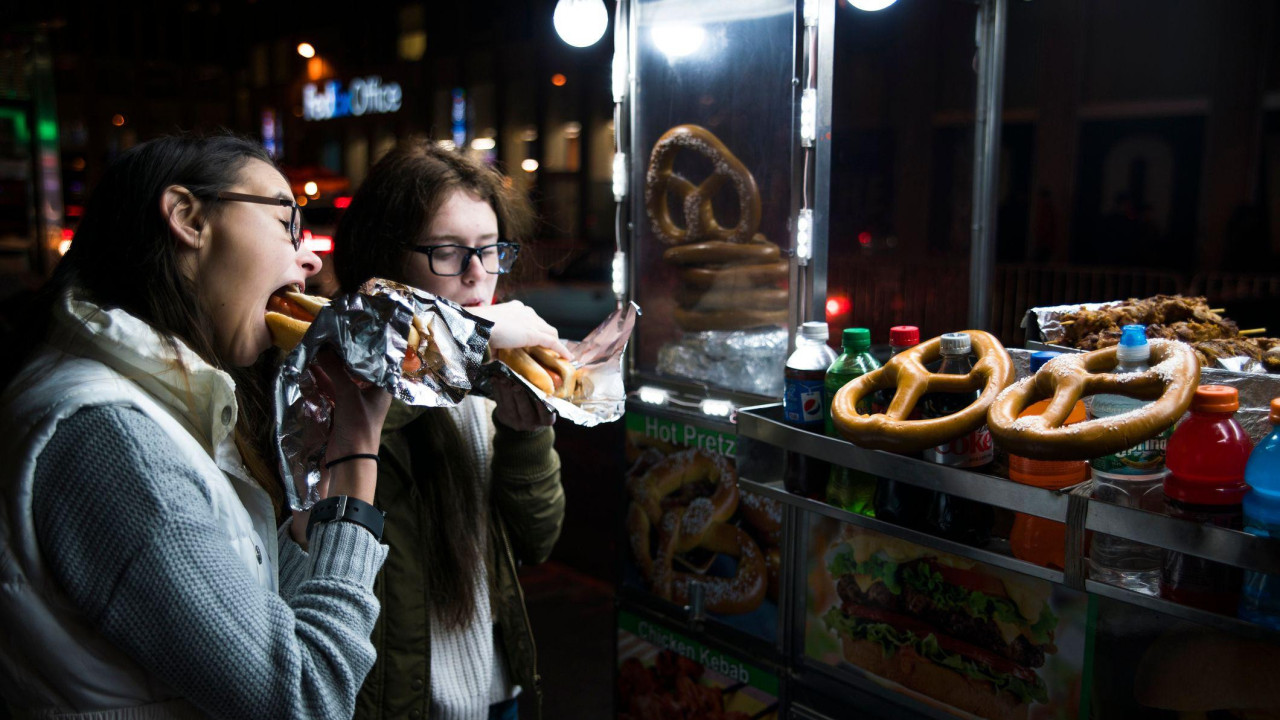 Two girls eat hot dogs outside a fast food stall. It is dark and they are both wearing jackets and jumpers.