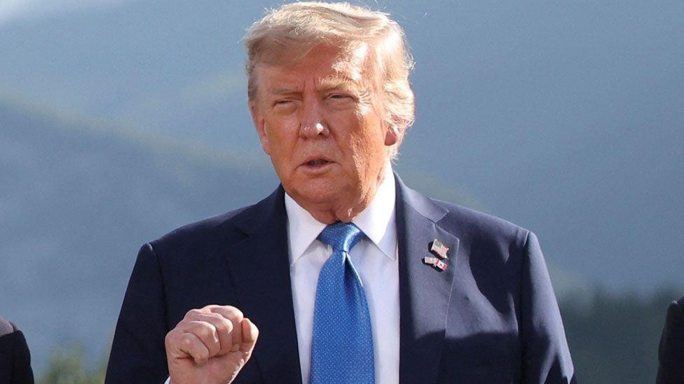 Donald Tramp napolju, u beloj košulji i plavom odelu stišće pesnicu dok priča na G7 u Kanadi