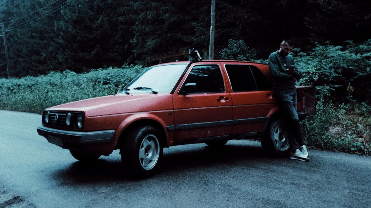 Muškarac naslonjen na crveni automobil koji je preprečio put, gleda u telefon